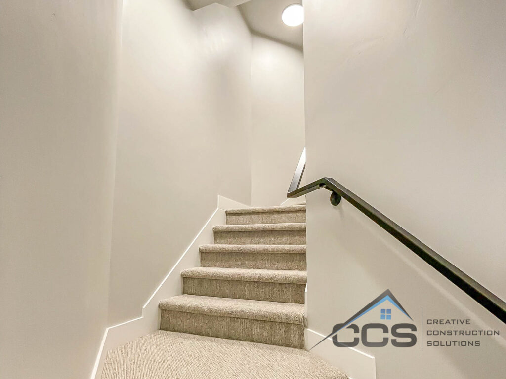 An image of a basement staircase looking up from the bottom. The stairs curve up and to the right. A railing is on the right side. A logo for Creative Construction Solutions, CCS of Utah is on the bottom right-hand corner.
