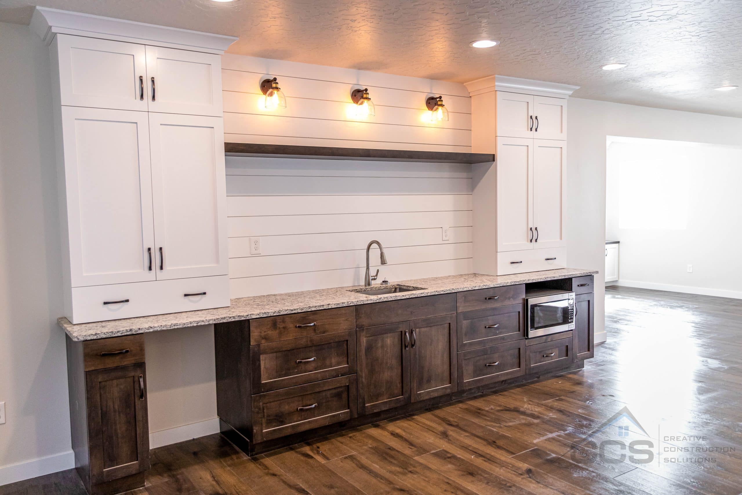 An image of a modern finished basement wet bar. Features include white cabinetry on the top with matching white clapboard. Dark oak stained cabinets below with matching floating shelves above sink. Granite countertops.