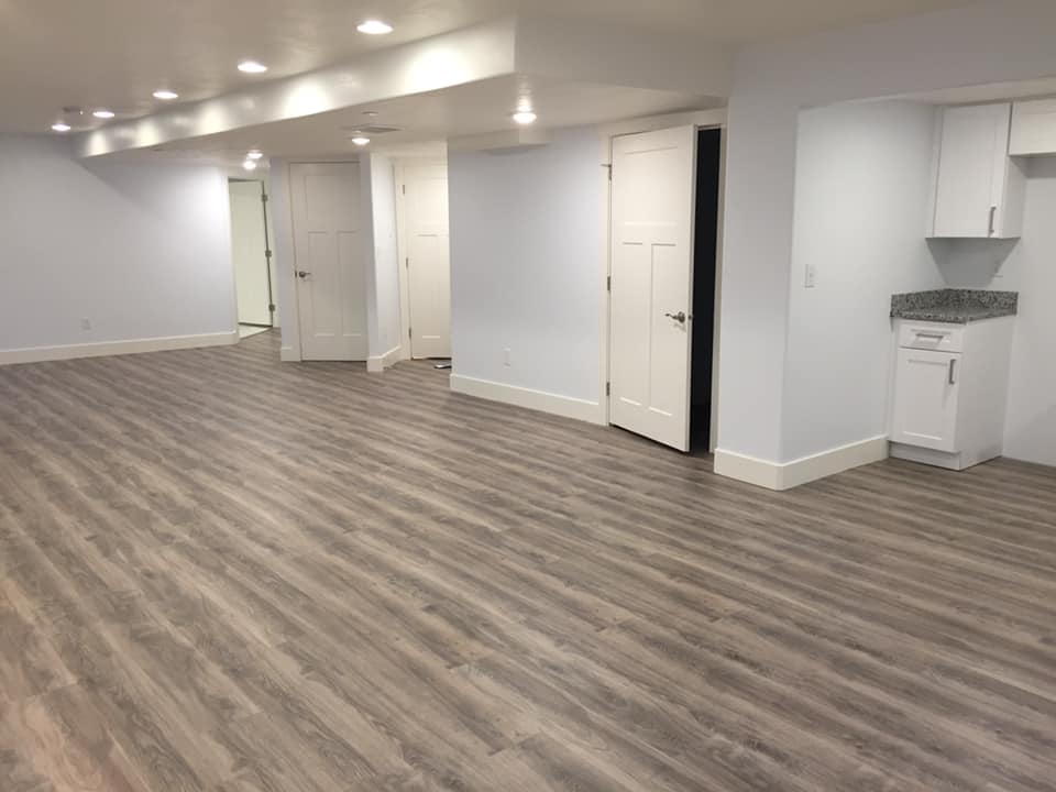An empty basement room with wooden flooring, white walls, doors, and a small cabinet.
